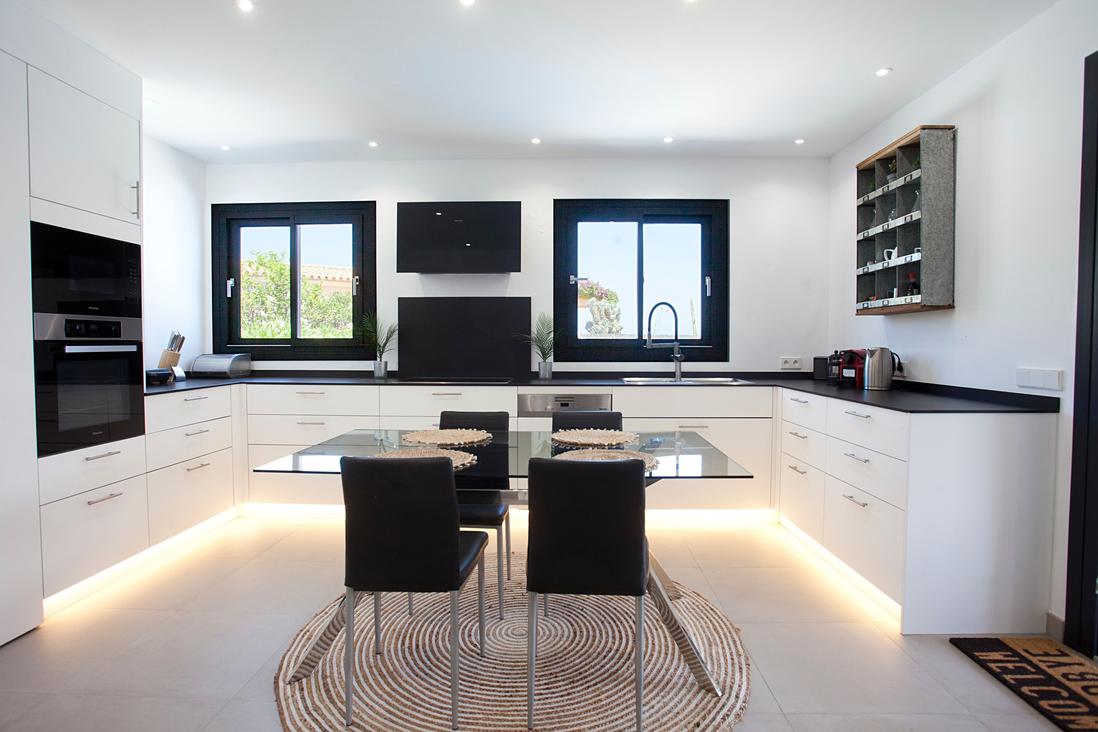 A modern kitchen with a central island, two black chairs, a glass dining table, and a built-in TV above the island.