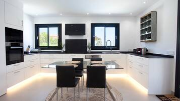 A modern kitchen with a central island, two black chairs, a glass dining table, and a built-in TV above the island.