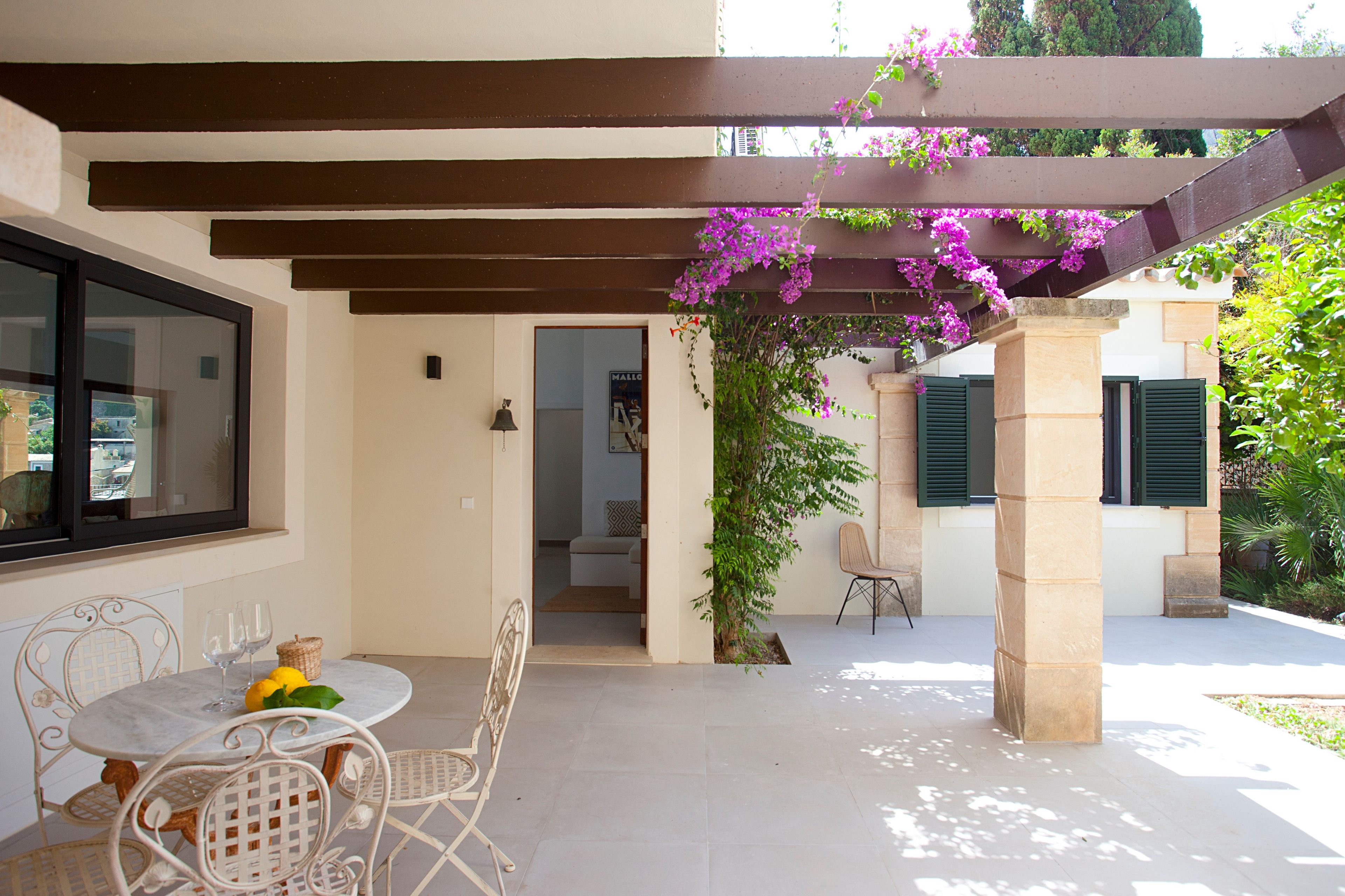 Un patio techado con una mesa y sillas, vistas del interior y una pérgola adornada con plantas florecientes.