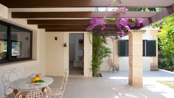 Un patio techado con una mesa y sillas, vistas del interior y una pérgola adornada con plantas florecientes.