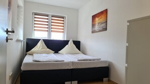 Een slaapkamer met een bed, twee ramen met jaloezieën, een schilderij aan de muur en een houten vloer.