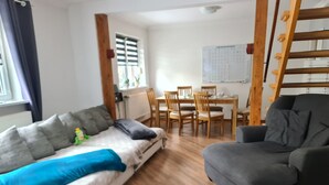 Een kamer met een bank, een stoel, een eettafel met stoelen, een ladder en een raam met jaloezieën.
