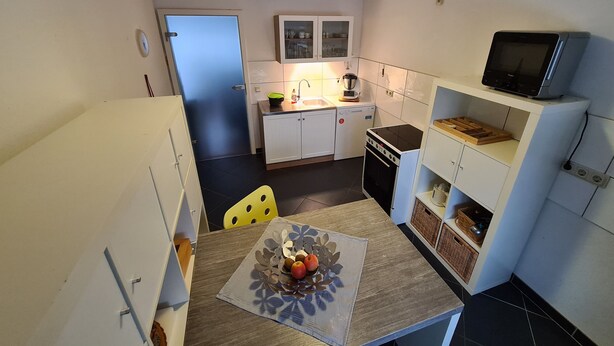 Een compacte keuken met witte kastjes, een magnetron, een fornuis en een kleine eethoek met een tafel en stoelen.