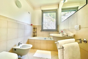 A bathroom with a bathtub, bidet, sink, and a window providing a view of greenery. - Gaeta 'lake View' (San Siro)