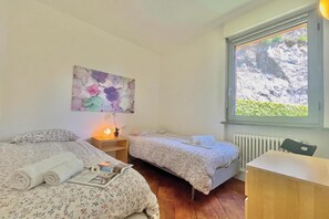 A bedroom with two beds, a bedside table with a lamp, a window with a view of a rocky landscape, and a wall-mounted artwork. - Gaeta 'lake View' (San Siro)