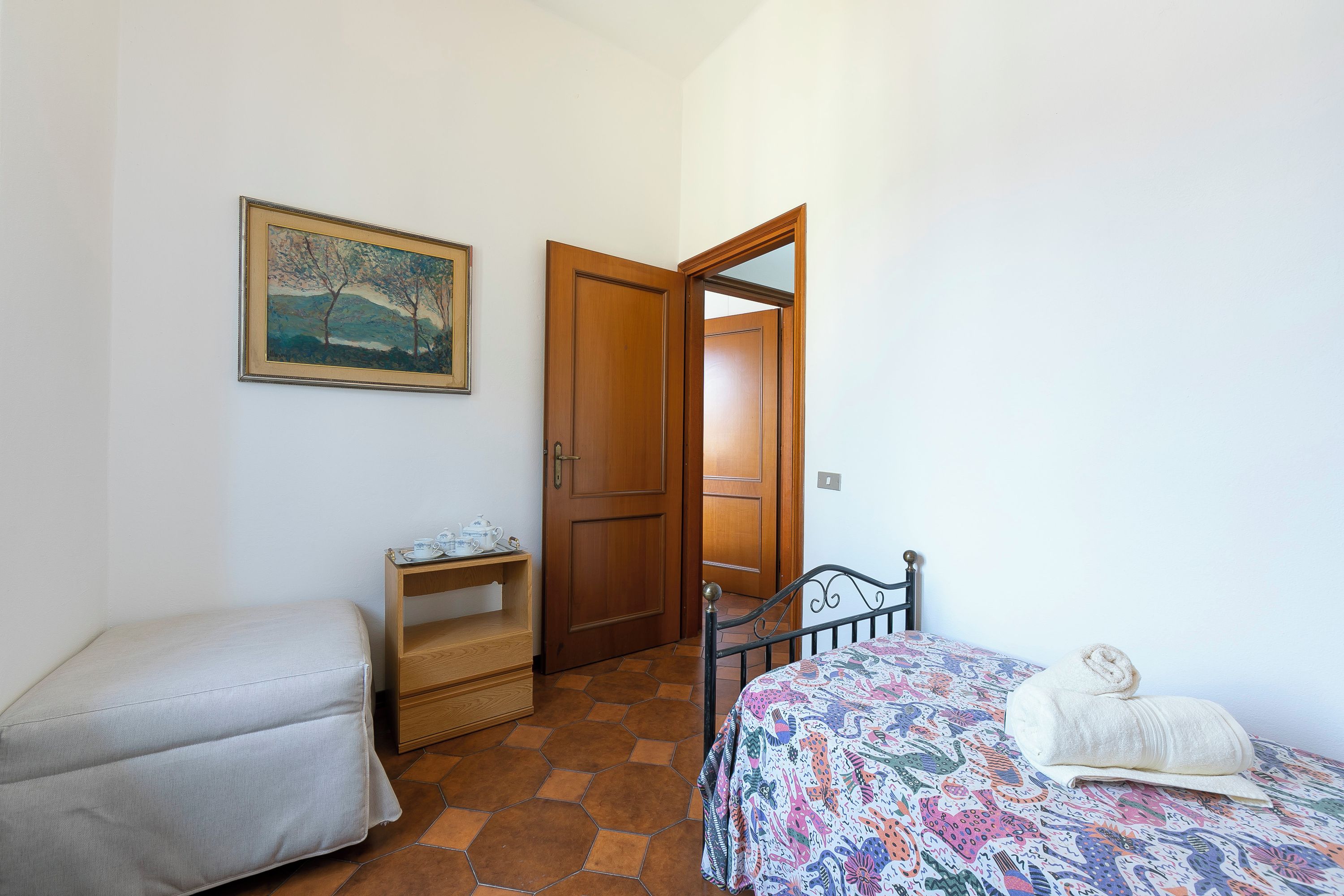 A bedroom with a bed, a bedside table, a chair, a door, and a painting on the wall.