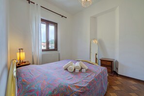 Ein Hotelzimmer mit einem Bett, einem Nachttisch mit Lampe, einem Fenster mit Vorhängen, einem Spiegel und einer kleinen Holzkommode.