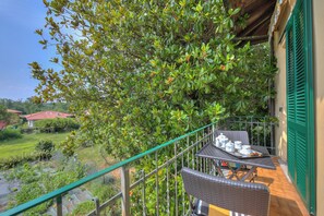 Ein Balkon mit Korbmöbeln, einem Tisch mit Teeservice und Blick auf einen Garten und Häuser.