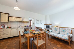A spacious living area with a wooden dining table, chairs, and a floral sofa. A kitchen with light-colored cabinets and a tiled backsplash is visible in the background.