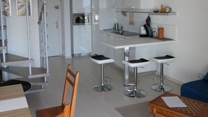 Ett modernt kök med vita skåp, en köksö och barstolar. I vardagsrummet finns ett matbord i trä med stolar och en blå soffa. En trappa med ett glasräcke leder upp till en övre våning.
