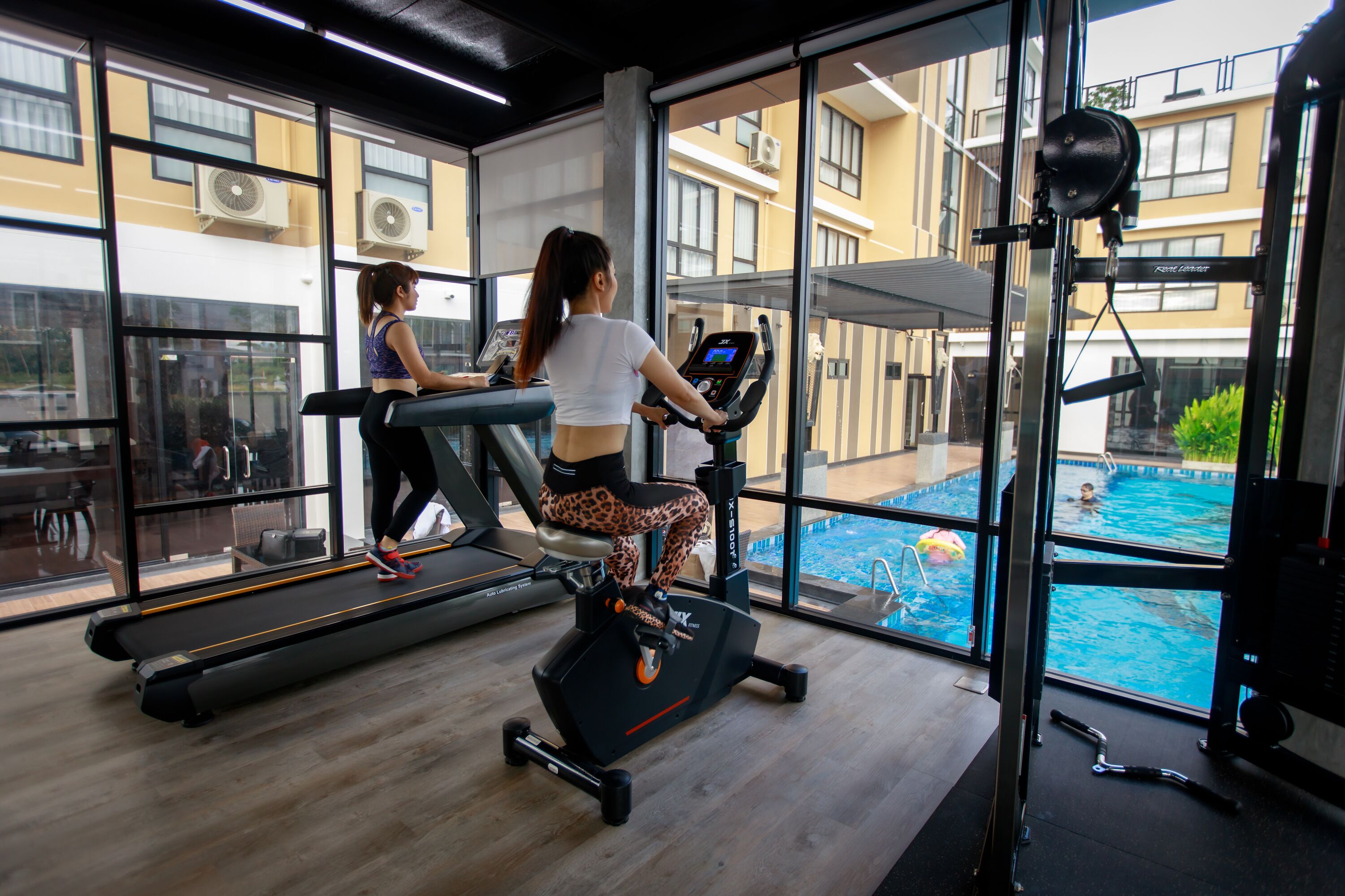 Un gimnasio con cintas de correr y bicicletas estáticas, amplias ventanas y vistas a una piscina.