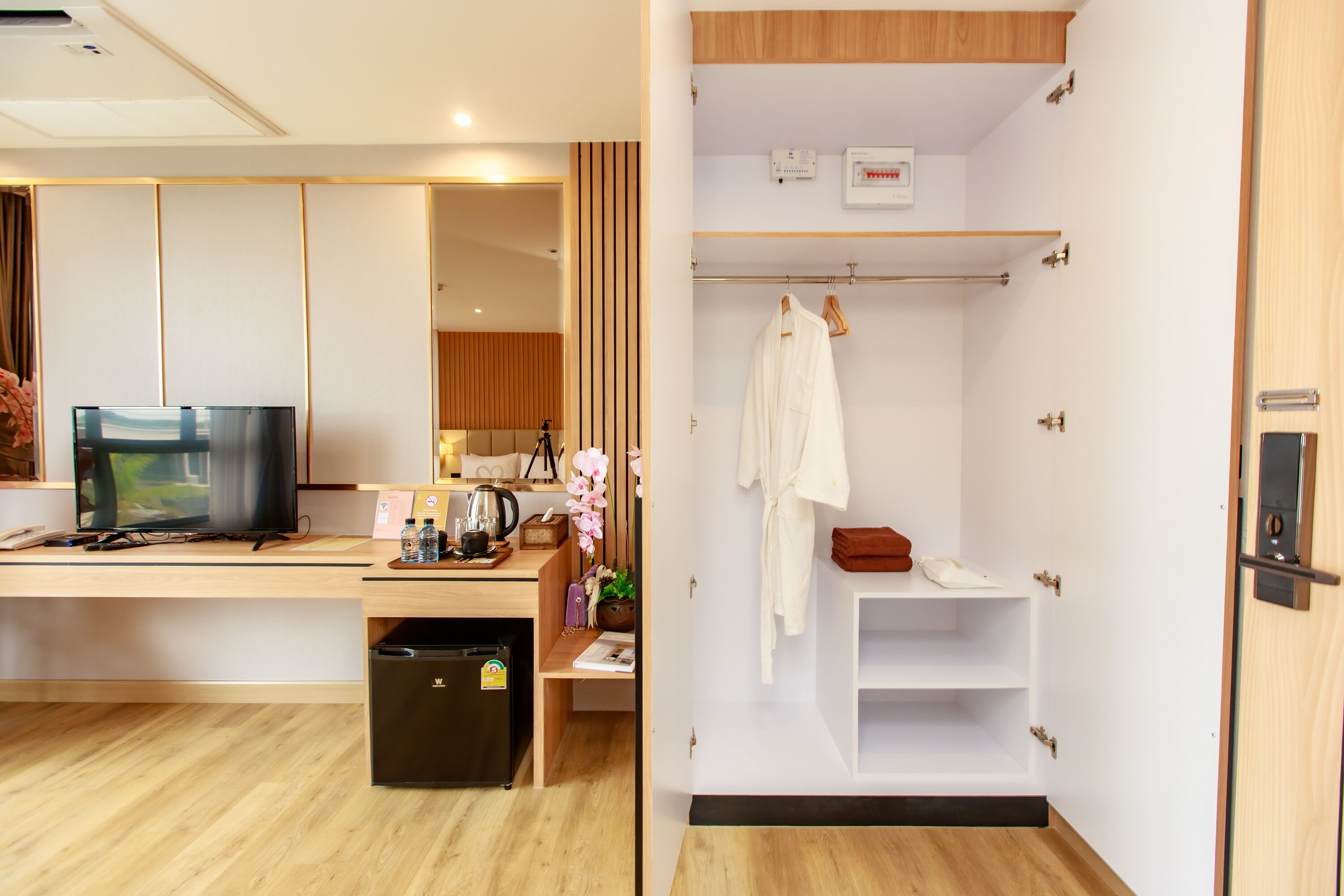 A hotel room with a wooden desk, a television, a minibar, a small refrigerator, a bathroom with a shower, a towel rack, and a shelf with toiletries.