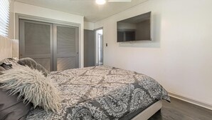 Un dormitori amb llit, ventilador de sostre, televisió penjada a la paret i una porta que condueix a una altra estança.
