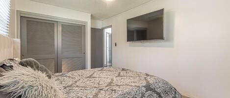 A bedroom with a bed, a ceiling fan, a TV mounted on the wall, and a door leading to another room.