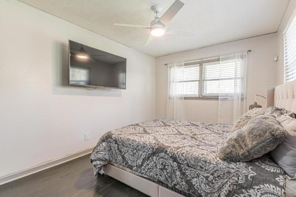 Une chambre à coucher avec un lit, un ventilateur de plafond, un téléviseur fixé au mur et une fenêtre avec des stores.