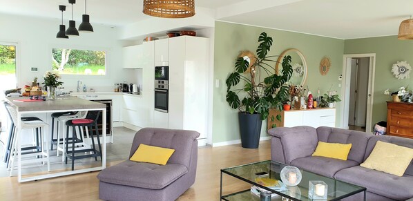 Una cocina moderna con armarios blancos, una zona de comedor con taburetes de bar, una planta grande en maceta y una sala de estar con sofá y mesa de centro.