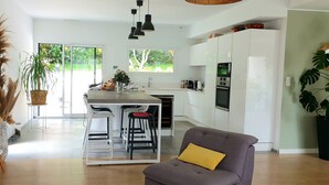 A modern kitchen with white cabinets, a central island, and bar stools. There is a dining area with a gray sofa and a TV on a wooden table. - Modern villa with all comforts (Saint-Génies-des-Mourgues)