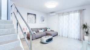 A modern living room with a grey sofa, a glass coffee table, and a staircase with glass railings.
