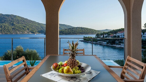 Uma mesa com uma cesta de frutas, duas cadeiras de madeira e vista para um lago e colinas.