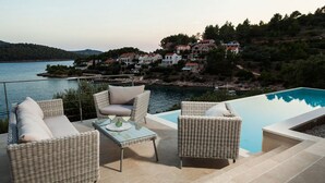 Área junto à piscina com mobiliário de vime e vista para uma colina com casas.