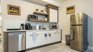 A modern kitchen with a stainless steel refrigerator, microwave, and dishwasher. There are two framed paintings on the walls and a small plant on the countertop. - The Premier Suite | 321Mbps WiFi | Perfect Stays (Airdrie)