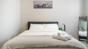 A neatly made bed with white linens and a folded towel, a cityscape painting on the wall, and a mirror on the right side. - The Premier Suite | 321Mbps WiFi | Perfect Stays (Airdrie)