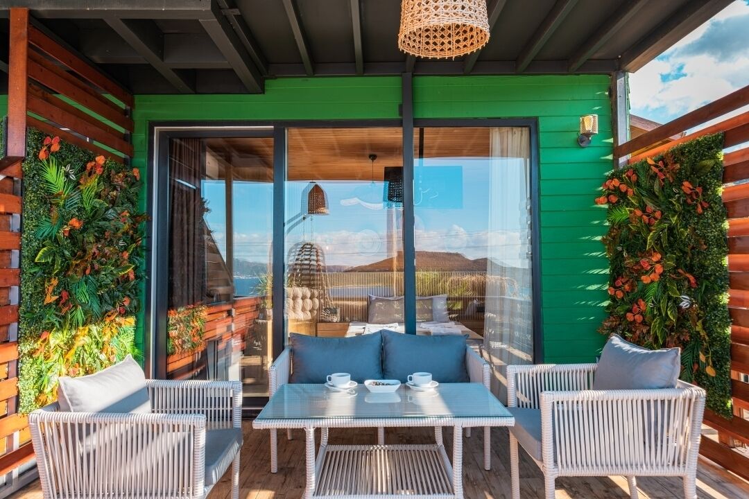 A modern outdoor seating area with wicker furniture and a glass table, surrounded by lush greenery and a view of the landscape.