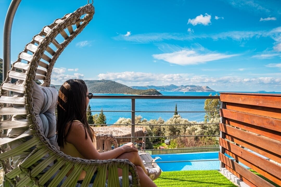 Une personne se détend dans un fauteuil en osier, avec vue sur une piscine et des montagnes.