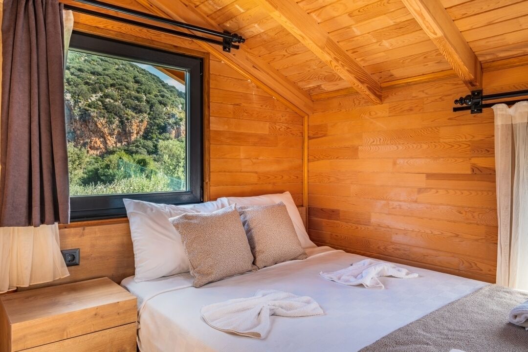 Ein Zimmer in einer Holzhütte mit einem Bett, einem Fenster mit Vorhängen und Blick auf einen bewaldeten Hang.