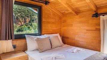 Ein Zimmer in einer Holzhütte mit einem Bett, einem Fenster mit Vorhängen und Blick auf einen bewaldeten Hang.