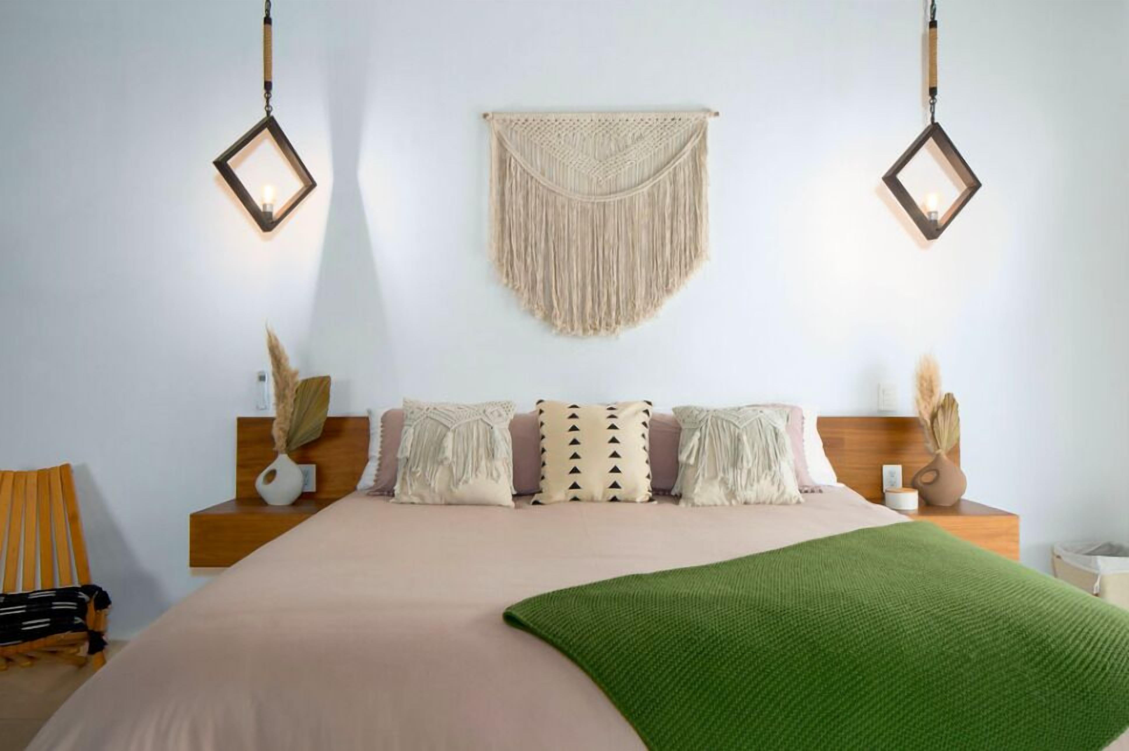A bedroom with a bed, two bedside tables, a hanging macrame wall hanging, and two wall-mounted lanterns.