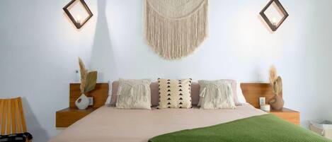 A bedroom with a bed, two bedside tables, a hanging macrame wall hanging, and two wall-mounted lanterns.