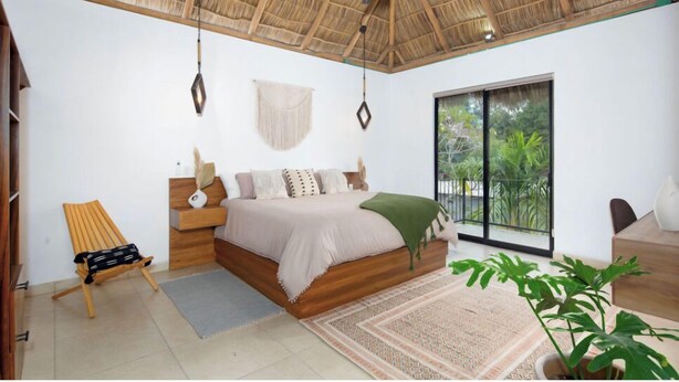 Ein Schlafzimmer mit einem Bett, einem Sessel, einer Pflanze und Blick auf Grün durch ein großes Fenster.