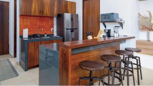 Eine moderne Küche mit Holzschränken, einer zentralen Kochinsel mit Hockern, einem Kühlschrank, einer Mikrowelle und einer Kaffeemaschine.
