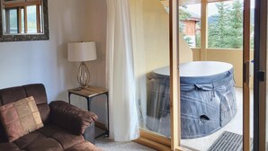 A room with a brown sofa, a small table with a lamp, and a view of a hot tub through a glass door. - Snow Creek Village #49 (Sun Peaks)