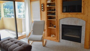 A living room with a fireplace, a chair, a shelf with decorative items, and a view of the outdoors. - Snow Creek Village #49 (Sun Peaks)