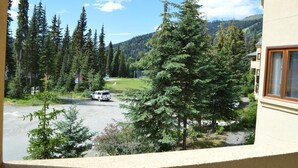 A view of a parking area with a white vehicle, surrounded by dense evergreen trees and a mountainous backdrop. - Snow Creek Village #49 (Sun Peaks)