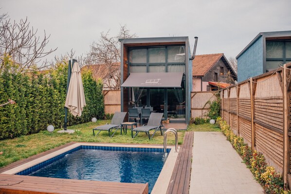 Un área de piscina exterior moderna con terraza de madera, sillones de descanso y un porche con paredes de vidrio.