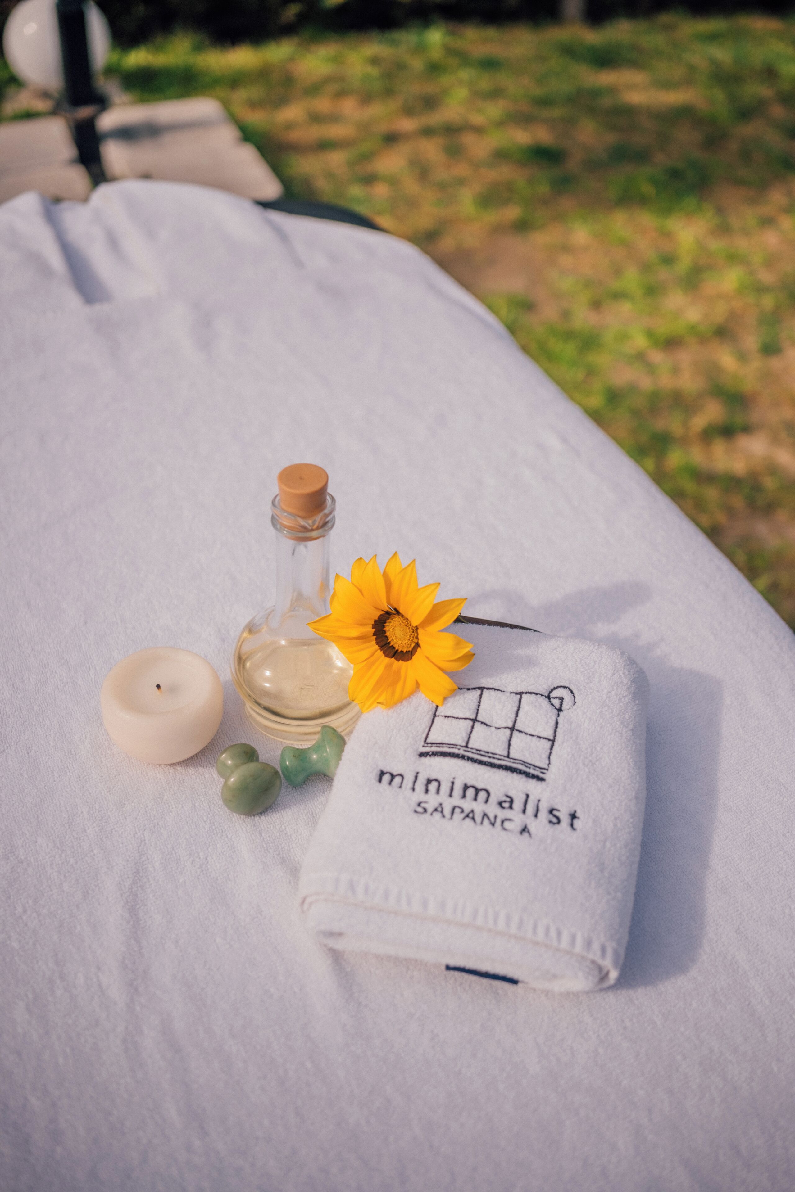 A towel with a sunflower and a bottle of oil, a candle, and a green soap on a white surface.