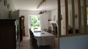 Een eetzaal met een lange tafel, stoelen en een kroonluchter. Op de muur hangt een klok en er is een raam met uitzicht op groen.