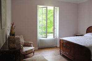 A bedroom with a wooden bed, a chair, a radiator, and a window with a view of greenery.