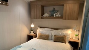 Cama bem arrumada com lençóis brancos e duas almofadas, mesa de cabeceira com abajur, e prateleira acima da cama com um enfeite de estrela e uma placa com a palavra 'Calm'.