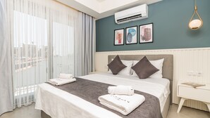 A modern hotel room with a large bed, white bedding, a grey headboard, and a view of the city through sheer curtains. - Kas Evderhan (Kas)