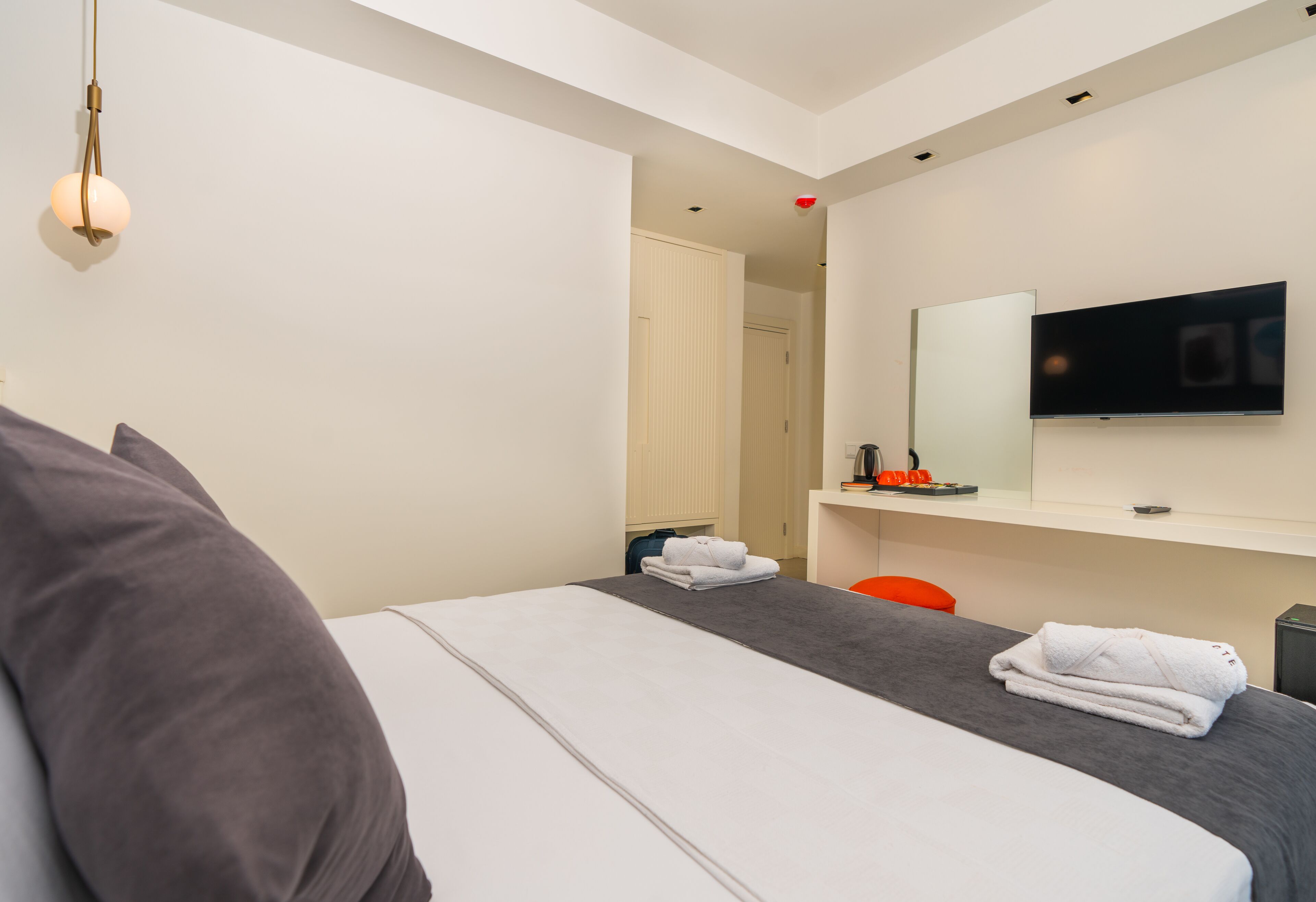 A modern hotel room with a bed, a TV, a mirror, and a small desk area.