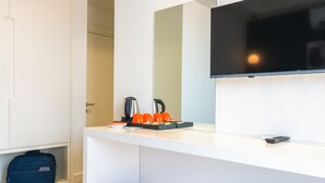 A modern hotel room with a white minibar, a flat-screen TV mounted on the wall, a small orange stool, and a blue suitcase on the floor. - Kas Evderhan (Kas)