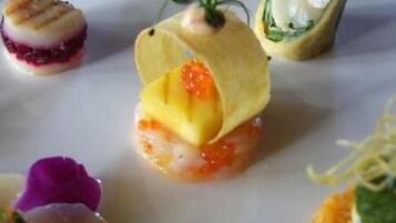 Verschiedene Gourmetgerichte auf einem weißen Teller, darunter ein gebratener Scampi, ein Sushi-Rolle und ein Gericht mit gelbem Belag und roten Kaviaarbällchen.