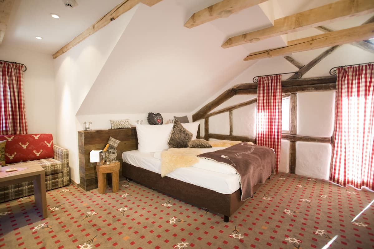 Un dormitorio con techo inclinado, cama con cabecera marrón, cortina a cuadros rojos y blancos y alfombra estampada.