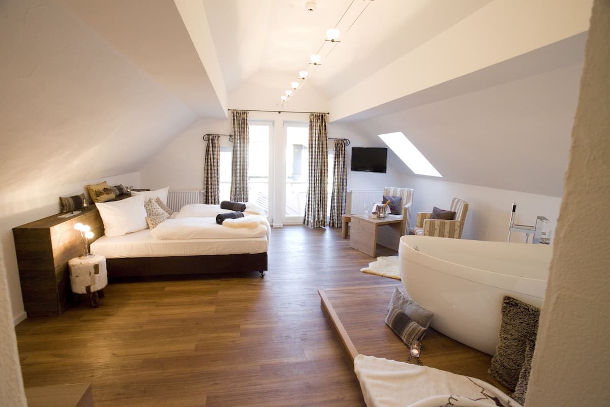 Un espacioso dormitorio en el ático con una cama grande, una zona de estar con un sofá blanco, suelo de madera y un tragaluz.