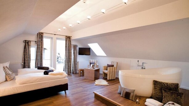 A modern bedroom with a large bed, a sitting area, a desk, a flat-screen TV, a large bathtub, and a skylight. - Alpin Lifestyle Hotel Löwen & Strauss (Oberstdorf)