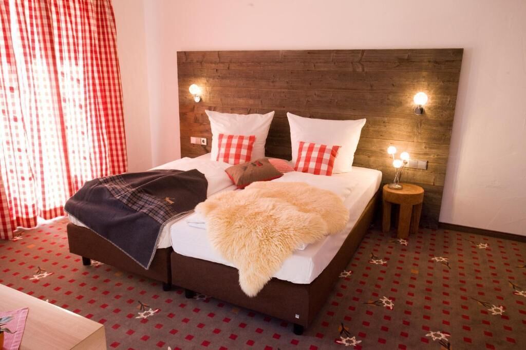 Ein Bett mit Holz-Kopfteil, weißer Bettwäsche und rot-weiß karierten Kissen. Zwei Nachttischlampen und ein kleiner Nachttisch mit einer Lampe sind vorhanden.